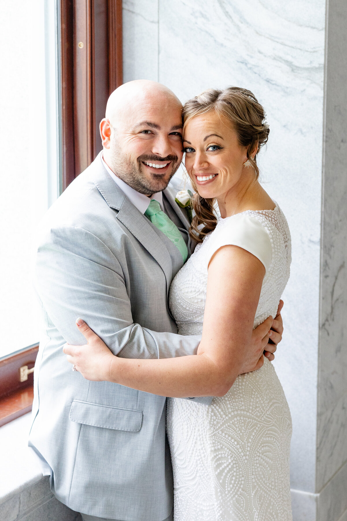 A smiling bride and groom embrace by a sunlit window, radiating happiness