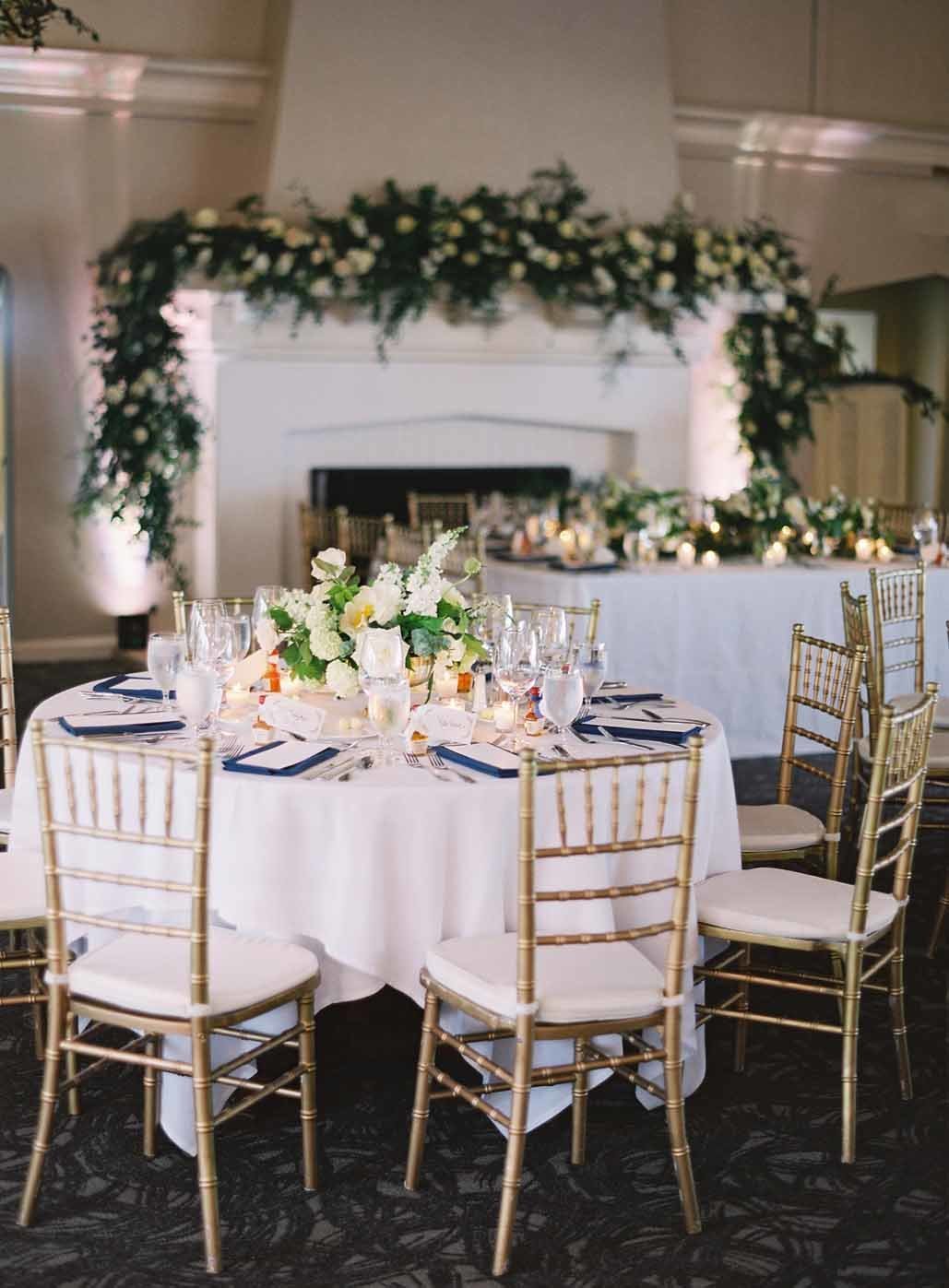 guest tables at Seattle Country Club wedding reception with ivory linens, blue napkins, gold Chivari chairs and greenery garland around fireplace behind head table