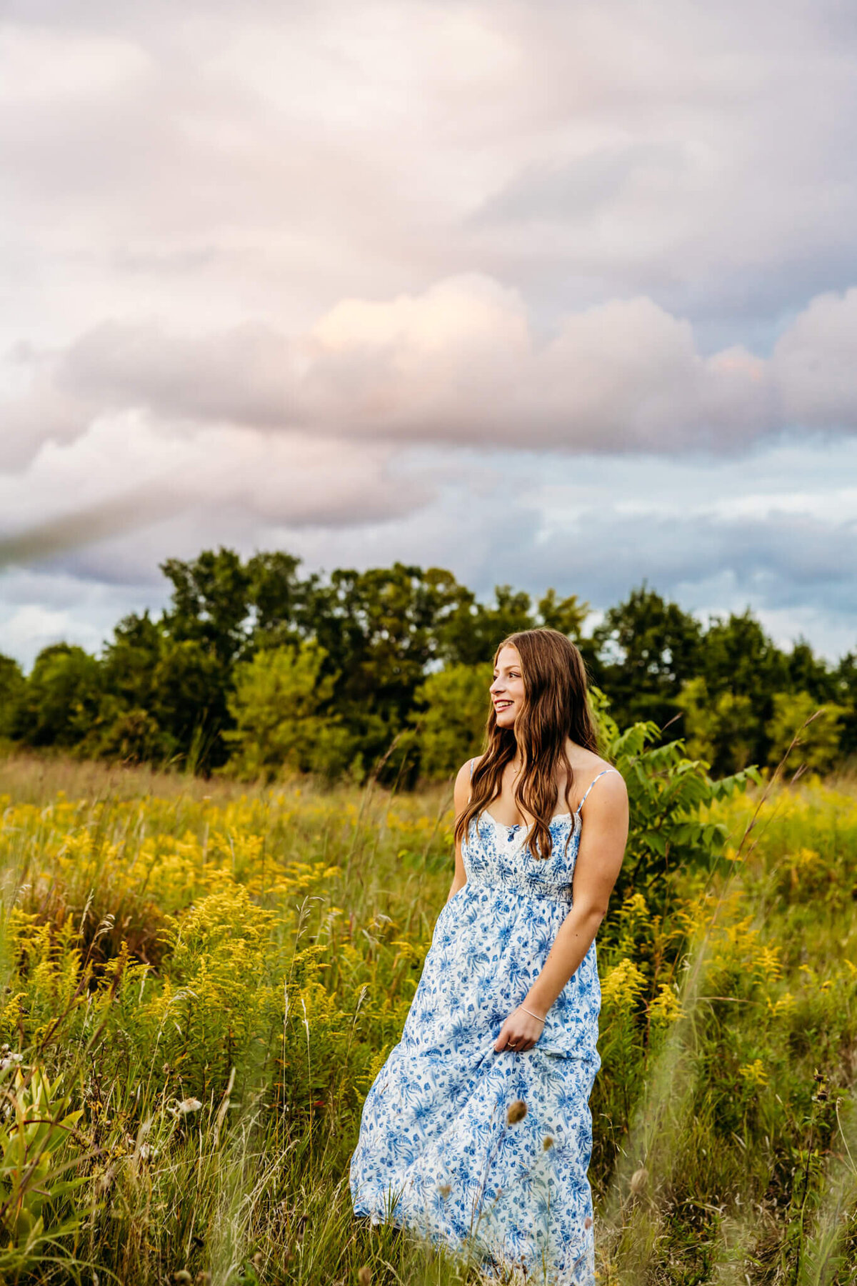 young lady in a blue and white patterned dress twirling in a flower field with a moody sky behind her captured by Ashley Kalbus.