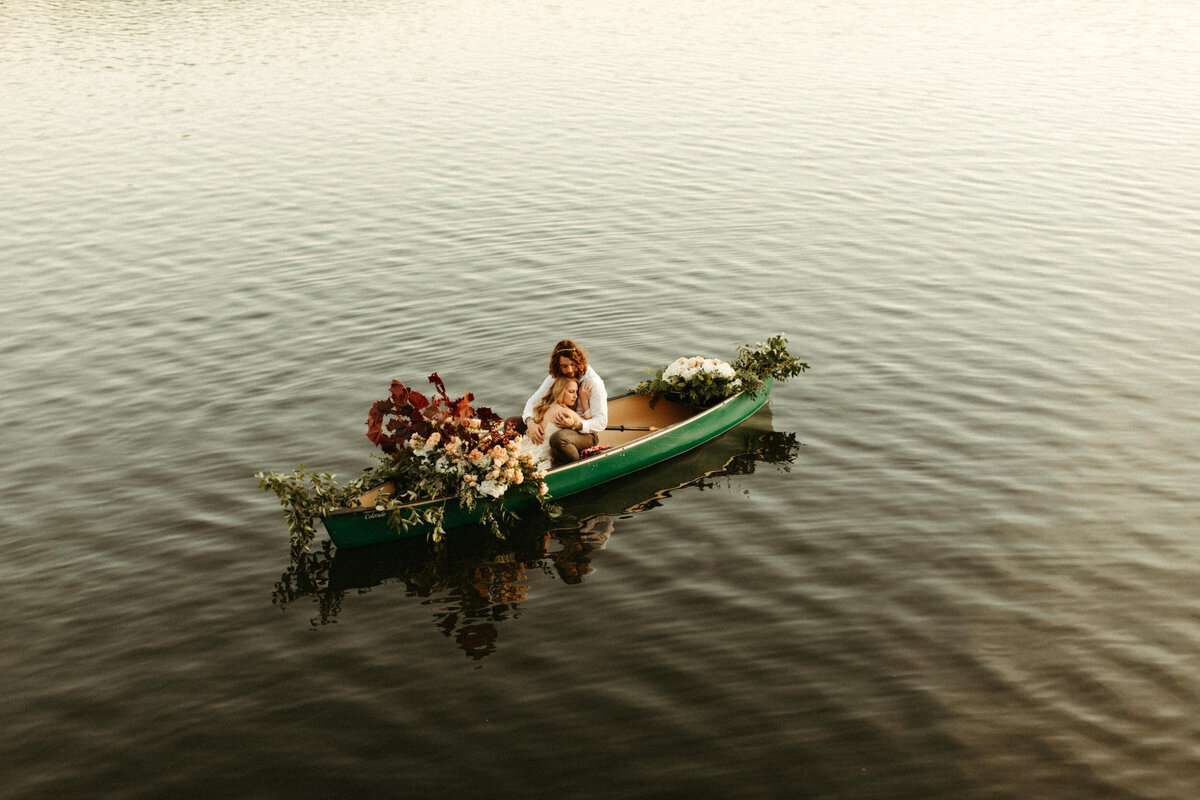 Groom embracing bride in canoe decorated with florals while they float in the middle of a lake at sunset