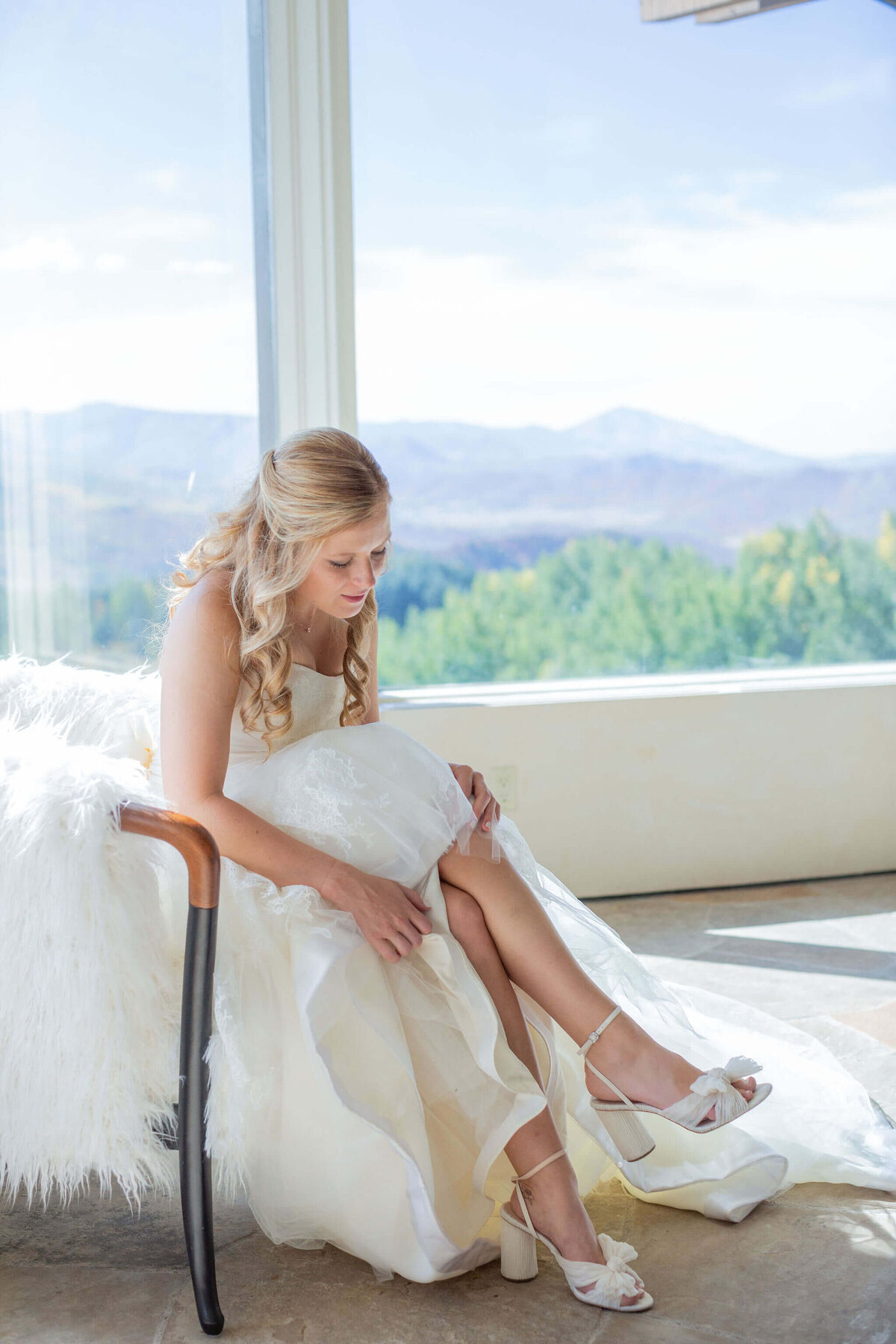 bride in white dress sits to buckle her white shoes