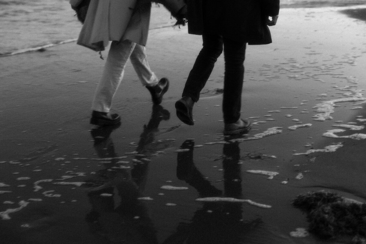 Couple walking holding hands with the waves splashing behind them on Newport Beach, Rhode Island, photographed by Andrea-Marie Photography