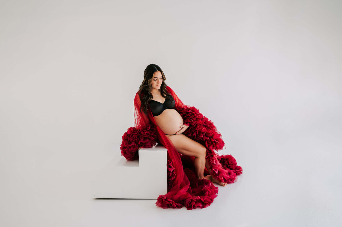 pregnant mom sitting on stairs in red robe looking at baby bump enjoying Joplin MO maternity photography