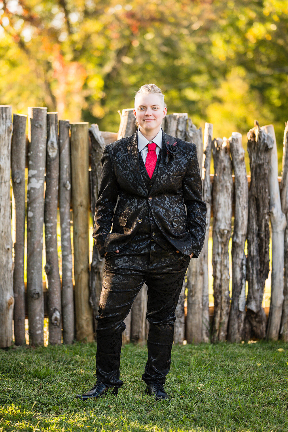A lesbian bride wearing a black floral textured suit and a red floral textured tie stands in the backyard of an Airbnb at golden hour in Roanoke, Virginia for a formal portrait.