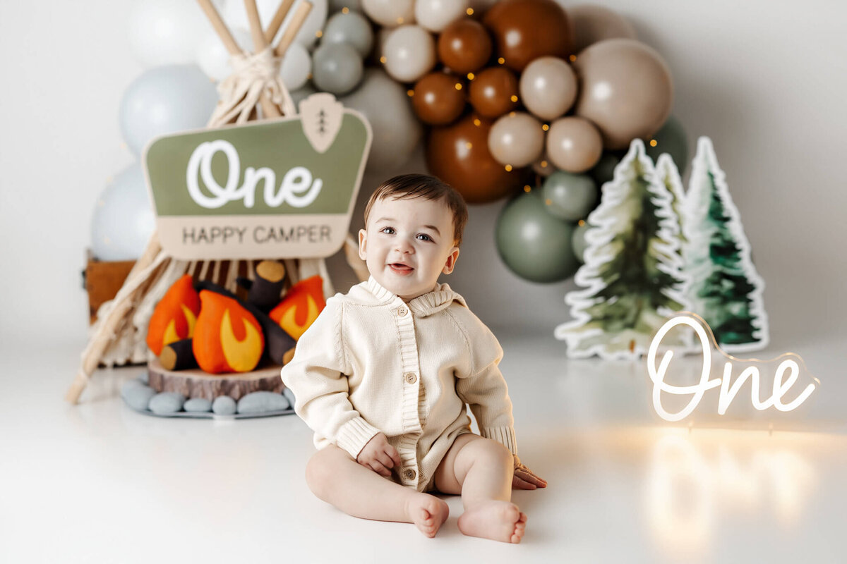 A baby wearing a cream sweater sits on the floor, smiling. Behind are balloon decor resembling a campfire and tent, with signs reading "One" and "Happy Camper." This cozy, camping-themed scene is perfect for families in Bloomington, Illinois seeking a memorable portrait photographer.