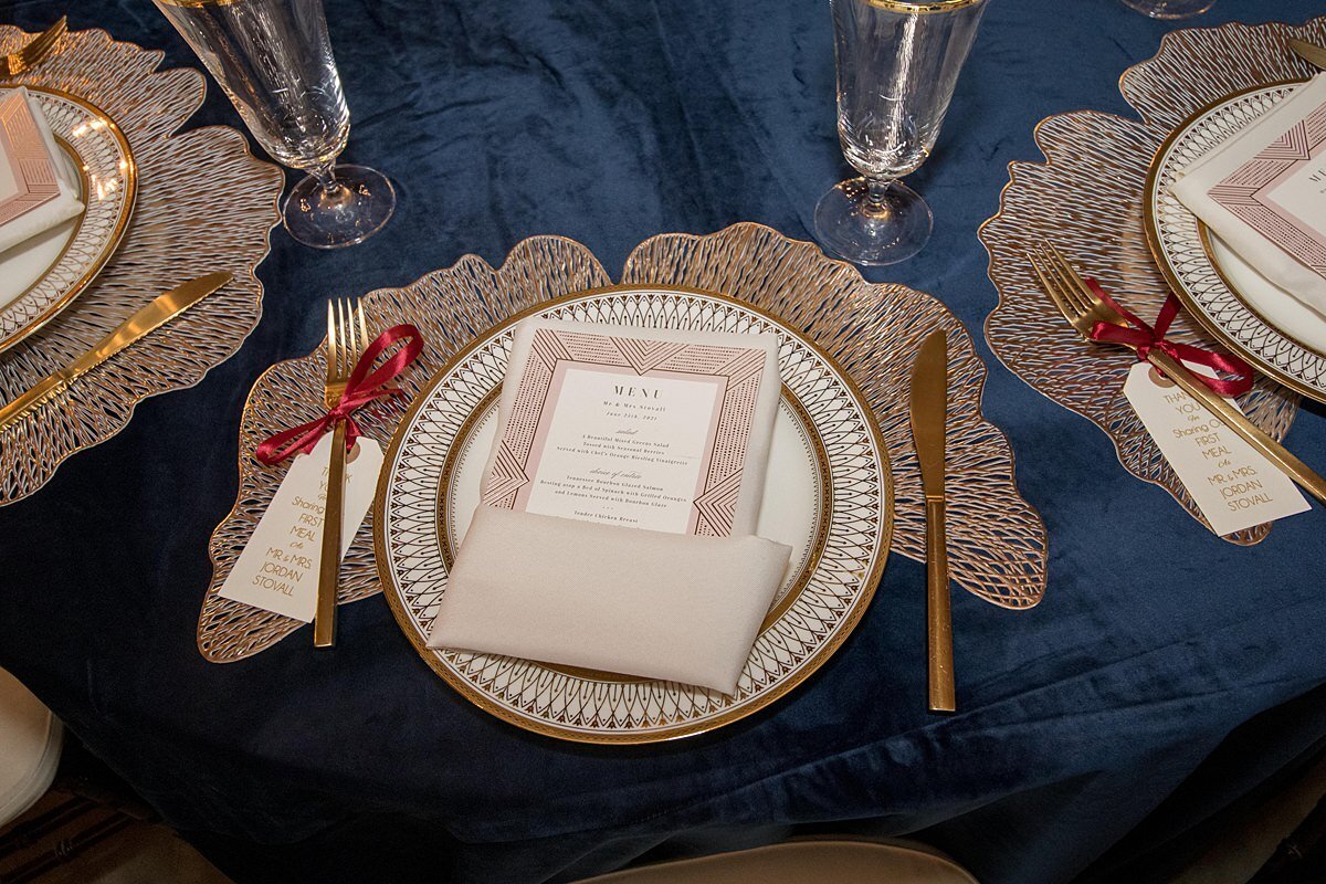 1920s themed wedding table setting of a ginko leaf charger with an ivory and gold plate set on top. A champagne linen napkin is wrapped around a menu card and the matte gold flatware has a thank you note tied to each fork with a burgundy ribbon. Footed crystal water goblets with a gold rim and a blue velvet tablecloth  give this reception a very decadent feel.