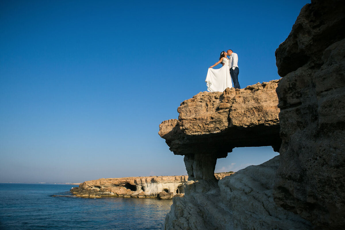 Bride & Groom standing on top of cliff overlooking the sea with bright blue sky