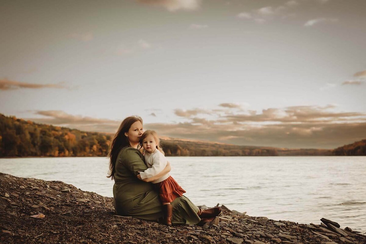 A mother sitting on a shore with her toddler daughter in her lap against her chest while they gaze into the distance