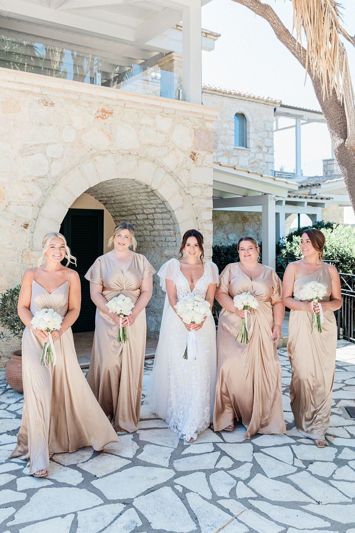 Bride with her bridesmaids in champagne satin dresses, holding white bouquets in a stone courtyard before a destination wedding ceremony