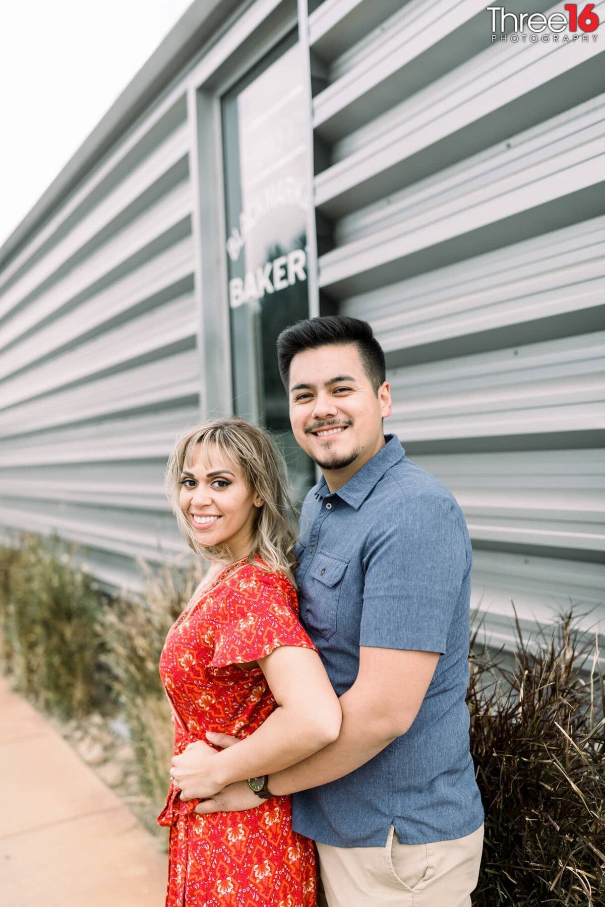 Groom to be holds his Bride from behind in front of a steel sided building