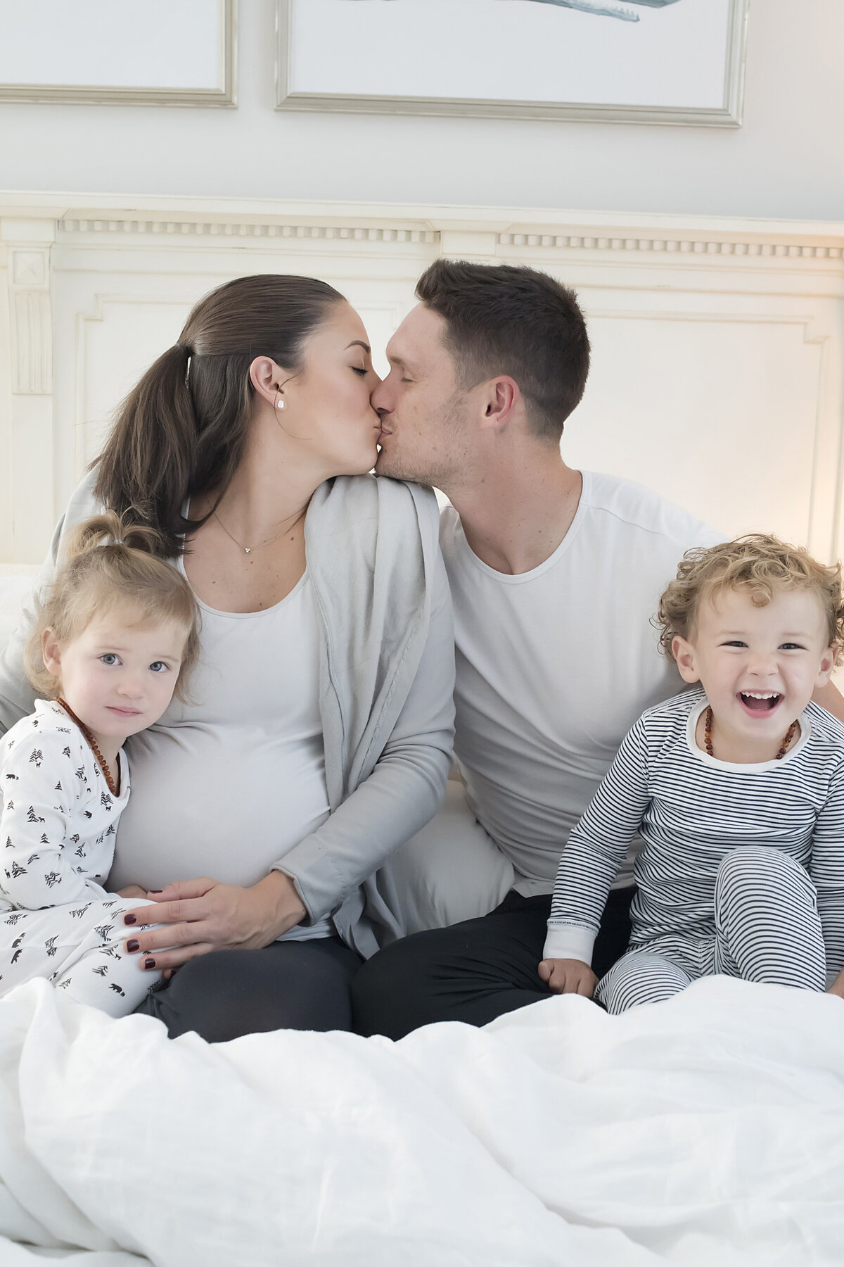 A pregnant couple sitting on a bed kissing with their two kids next to them 