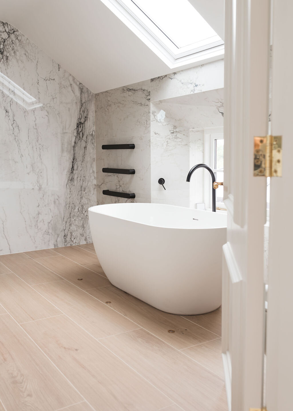 A modern white soaking tub in a grey marble bathroom with elegant black towel bars mounted to the marble walls and a skylight above.