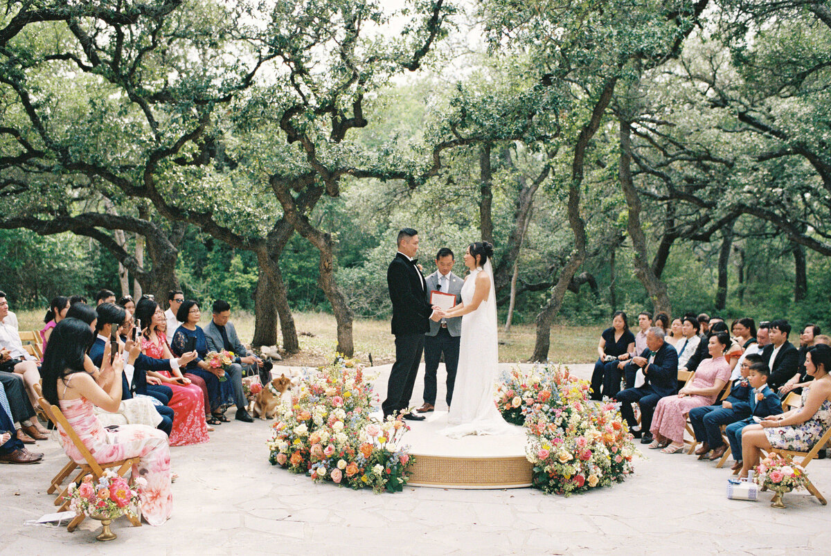 Outdoor wedding ceremony under oak trees with bride and groom exchanging vows surrounded by family and colorful floral arrangements