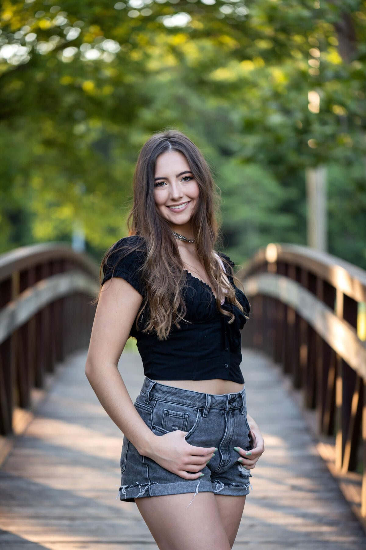 A pretty brunette senior girl poses with her thumbs in her pockets on walk bridge. Captured by senior photographer Dynae Levingston.