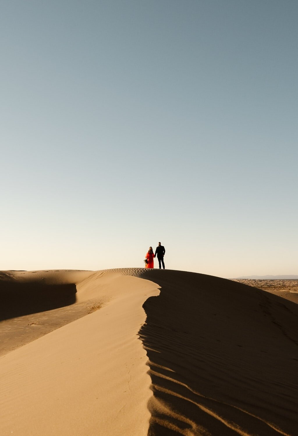 A couple stands on a ridge of the Glamis Sand Dunes, with the woman in a red dress and the man in dark attire, their silhouettes contrasting against the clear sky at dusk.