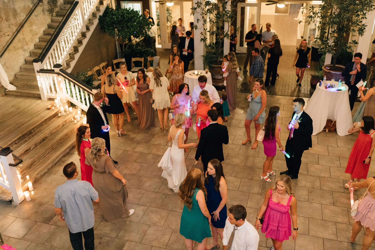 A group of people dressed in semi-formal attire dance and socialize at an outdoor wedding with candles, decorative plants, and a staircase in the background.