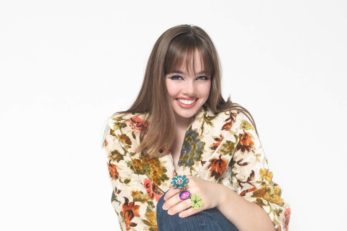 A smiling woman with long brown hair is wearing a floral-patterned blouse. She has colorful rings on her fingers and is sitting against a plain white background.