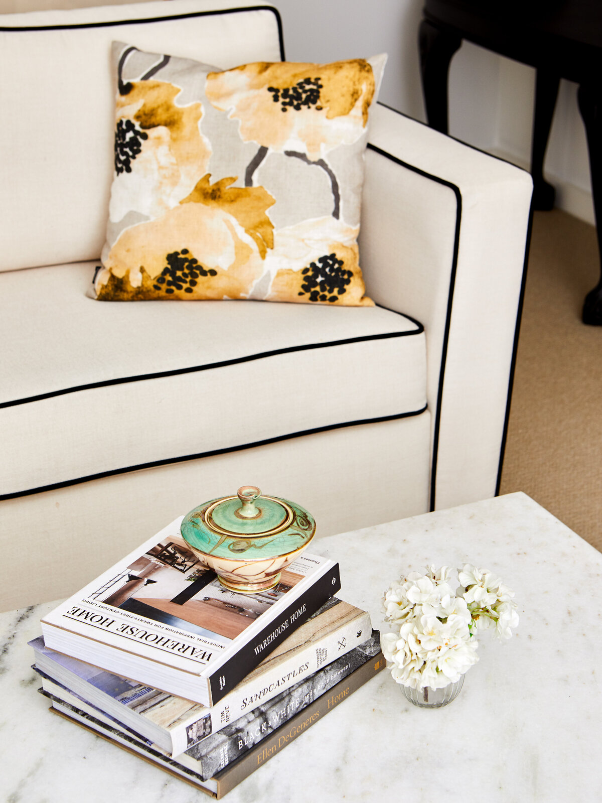 A detail shot of a white upholstered couch in a living room in Eynesbury, styled with a floral cushion in gold, black, and white. A marble coffee table is visible in the foreground with a stack of books.