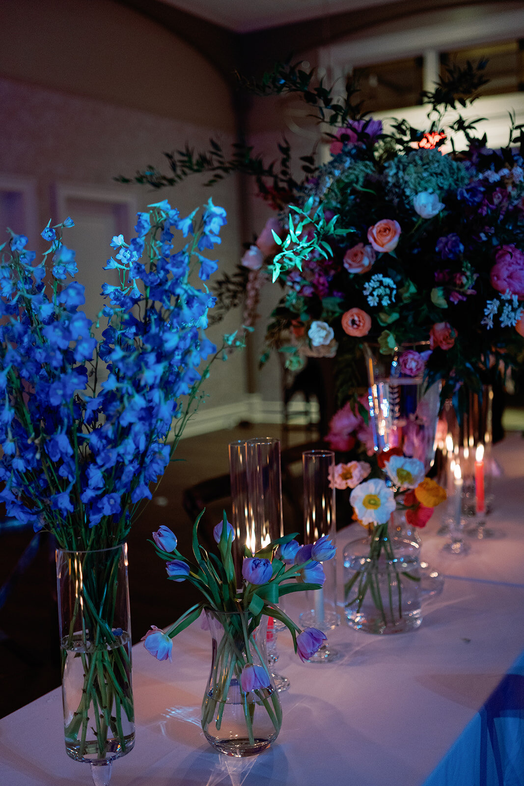 Vibrant reception table at Burritt on the Mountain featuring tall glass vases filled with vivid blue, pink, and orange blooms, surrounded by glowing candlelight.