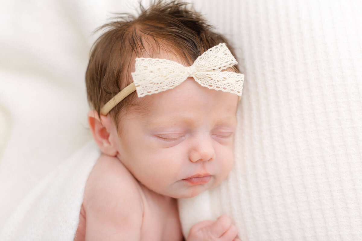 Newborn Baby wrapped in white and laying on a white textured blanket with a little white lace bow on her head. She has dark brown hair and is sleeping peacefully.
