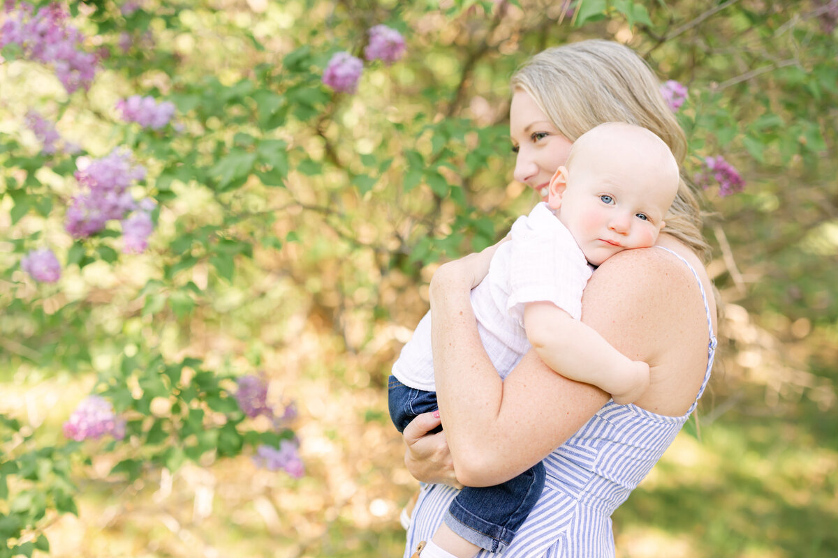 a mother standing in front of lilacs holds her baby closely as the baby lays into her shoulder