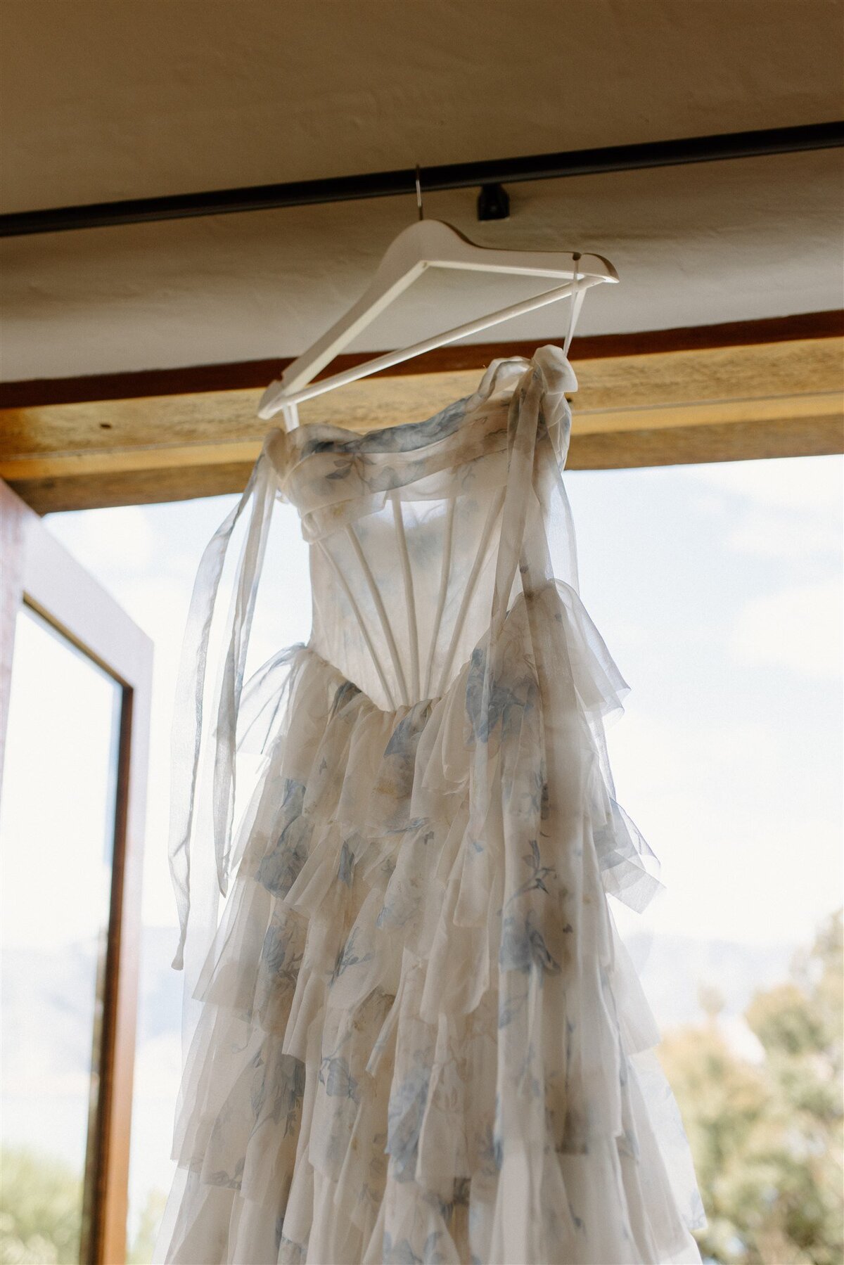 Light and airy floral wedding gown with layered ruffles and soft blue floral print, hanging by an open door with lake views outside.