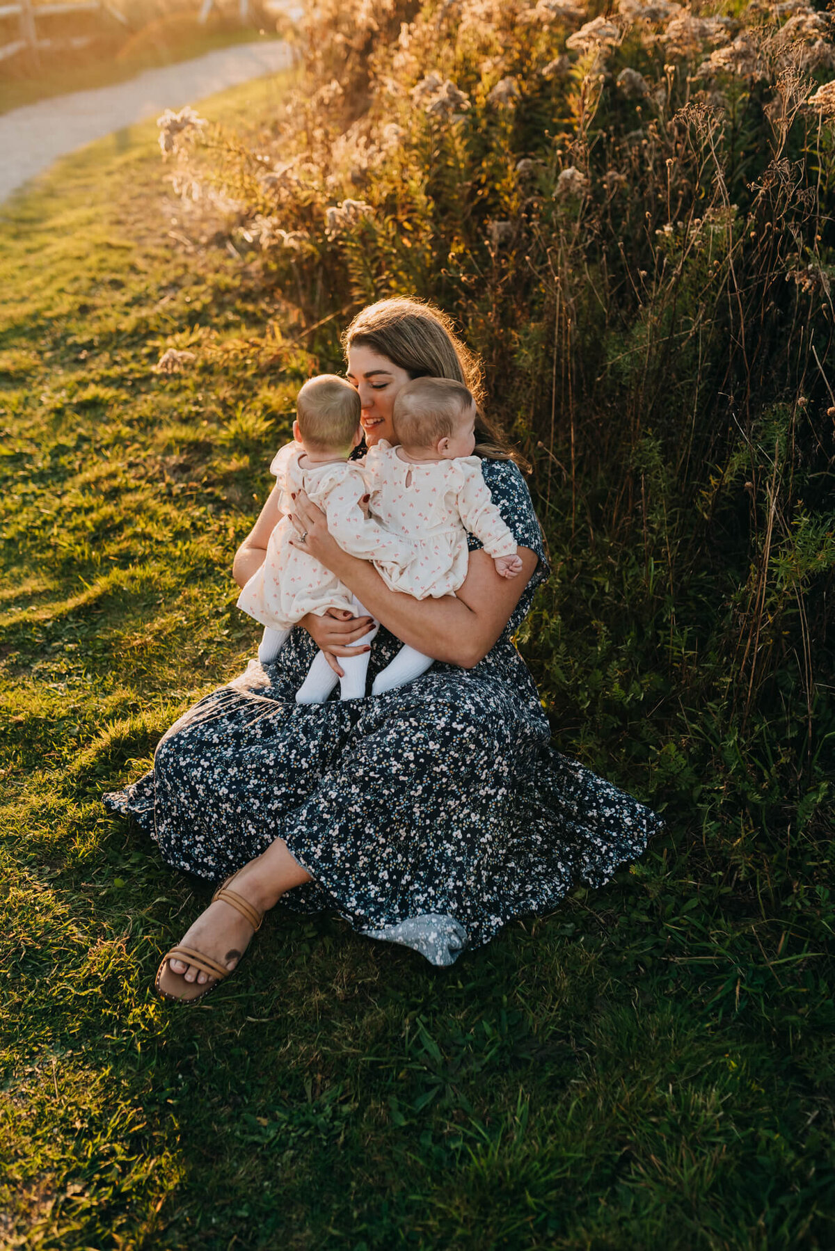 A mother holds her twin baby girls while sitting on the ground at sunset
