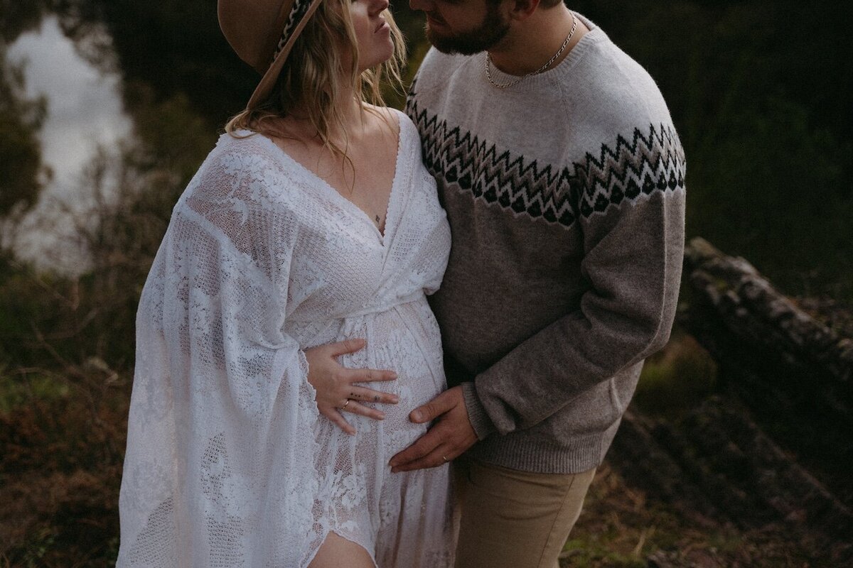 Couple s'échangeant un regard tendre, mains sur le ventre arrondi de la femme en robe blanche fluide. Photo en extérieur par Kamescop, photographe et vidéaste.