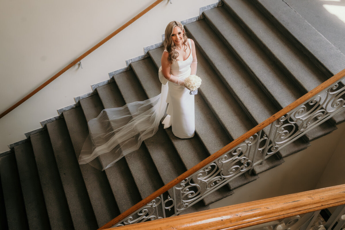 Auf einer großen Treppe stehend blickt die Braut nach oben in die Kamera.