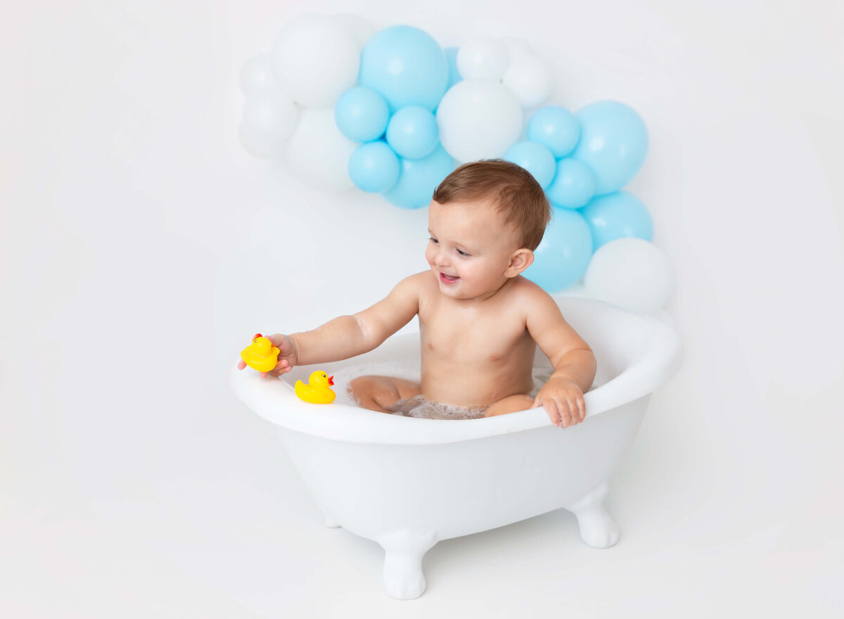 Baby boy sits in bubble bath white blue ballon garland cake smash