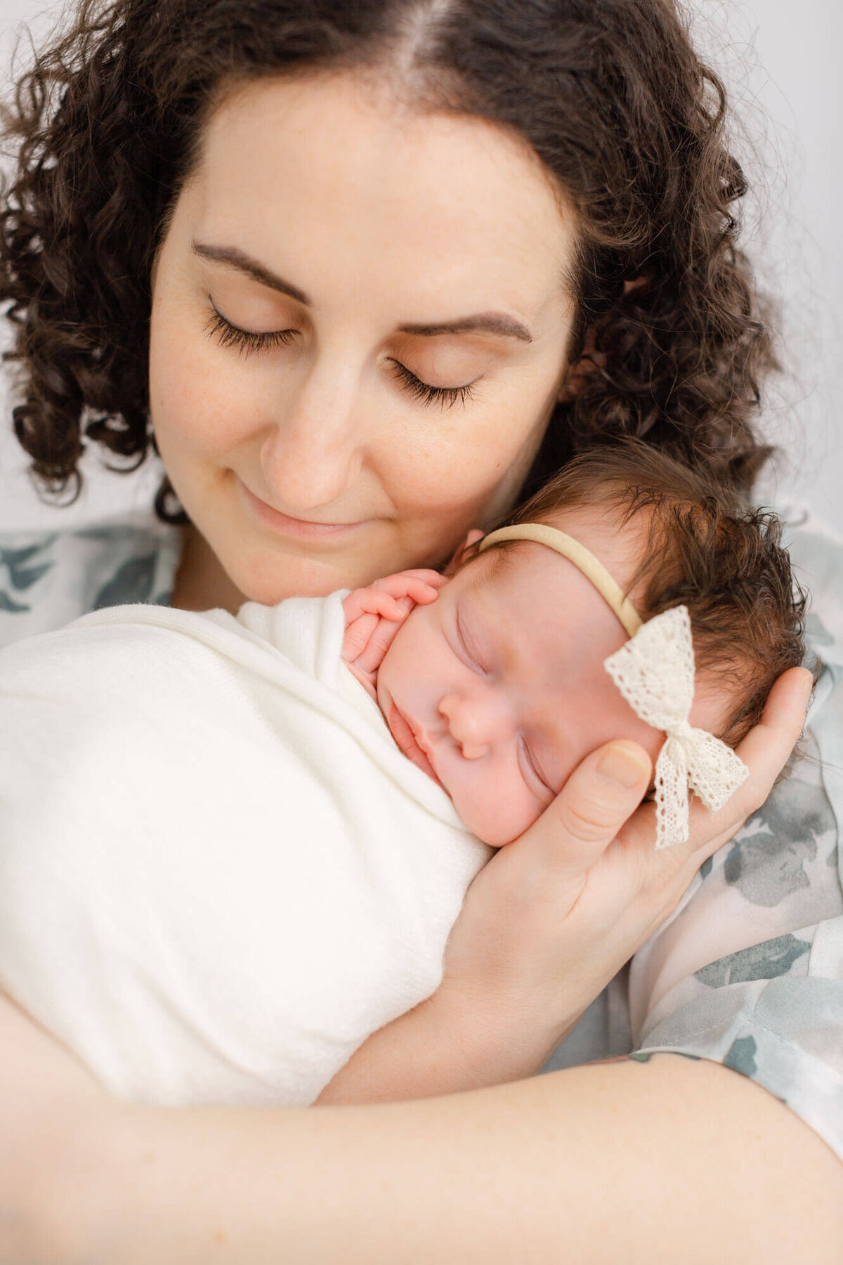 New mom with dark curly hair holding her baby up to her cheek. Mom has eyes closed and a small grin on her face. Baby is sleeping peacefully and wrapped up in a white blanket with her little hands out together resting on her cheek.