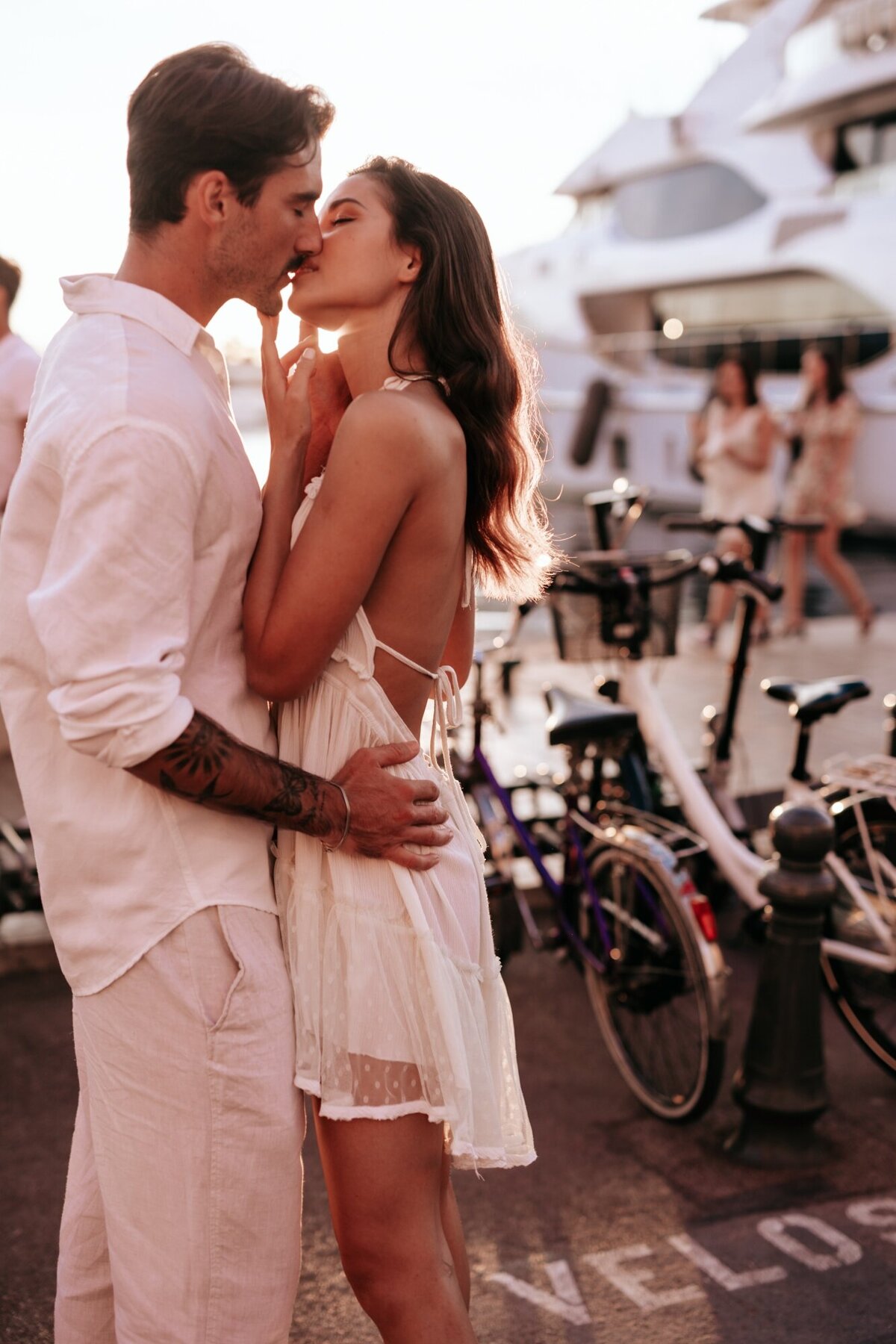 Couple sharing a kiss while his hand s are around her waist and hers are on his face with scooters and a yacht in the background.