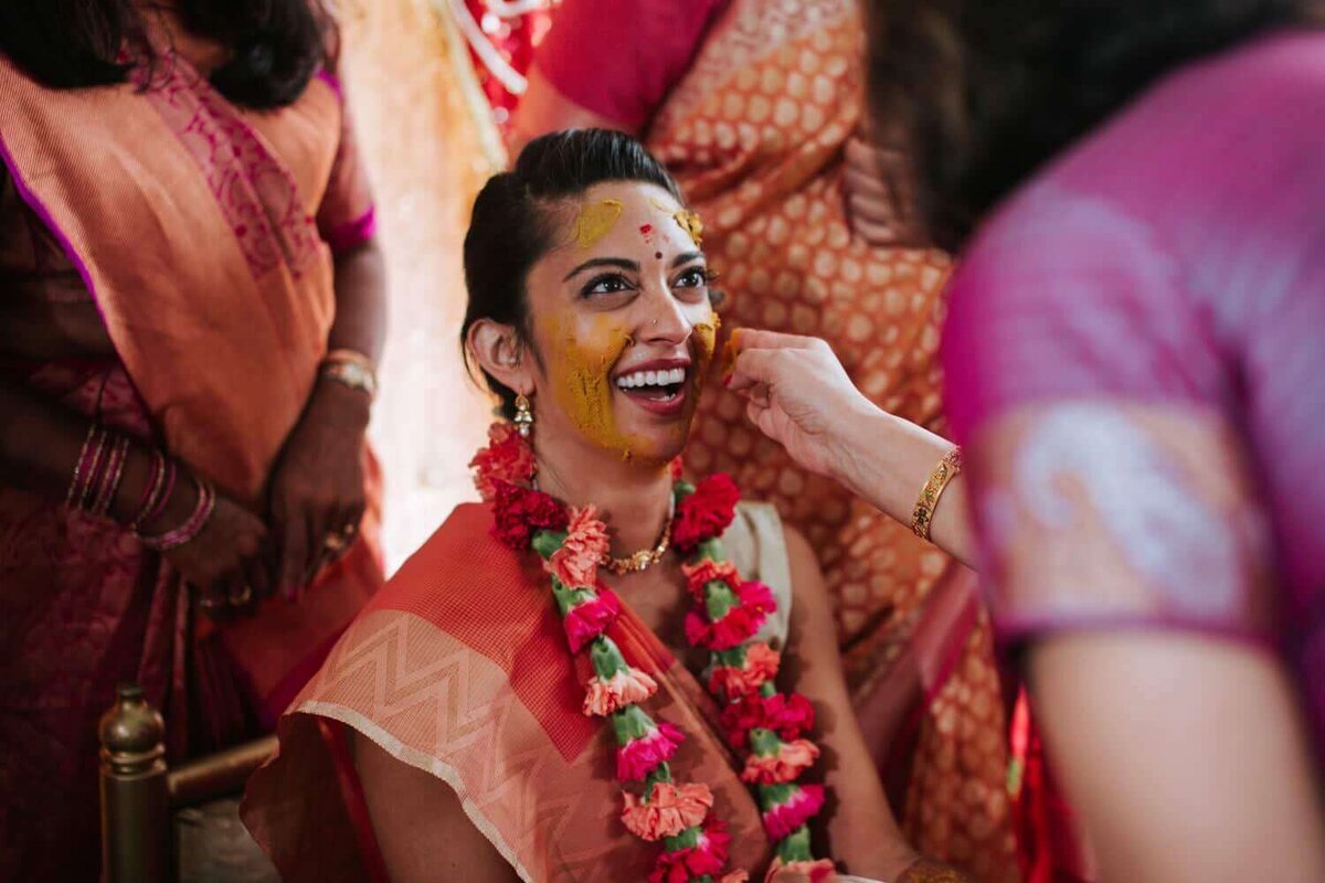 Bride at her Pithi/Haldi ceremony smiling as turmeric paste is being applied to her face.