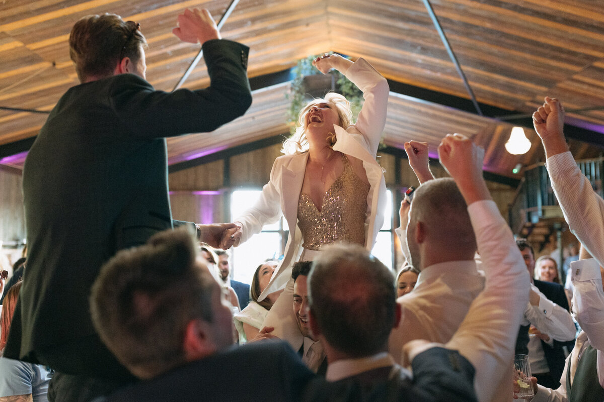 The bride, in a sparkling outfit and a white suit, is joyfully lifted into the air by her guests during the wedding celebration at Willow Marsh Farm. She smiles with excitement, her arms raised in the air as she is surrounded by guests cheering and dancing. The vibrant atmosphere of the reception hall, with purple lighting and a lively crowd, adds to the energy of this unforgettable moment.