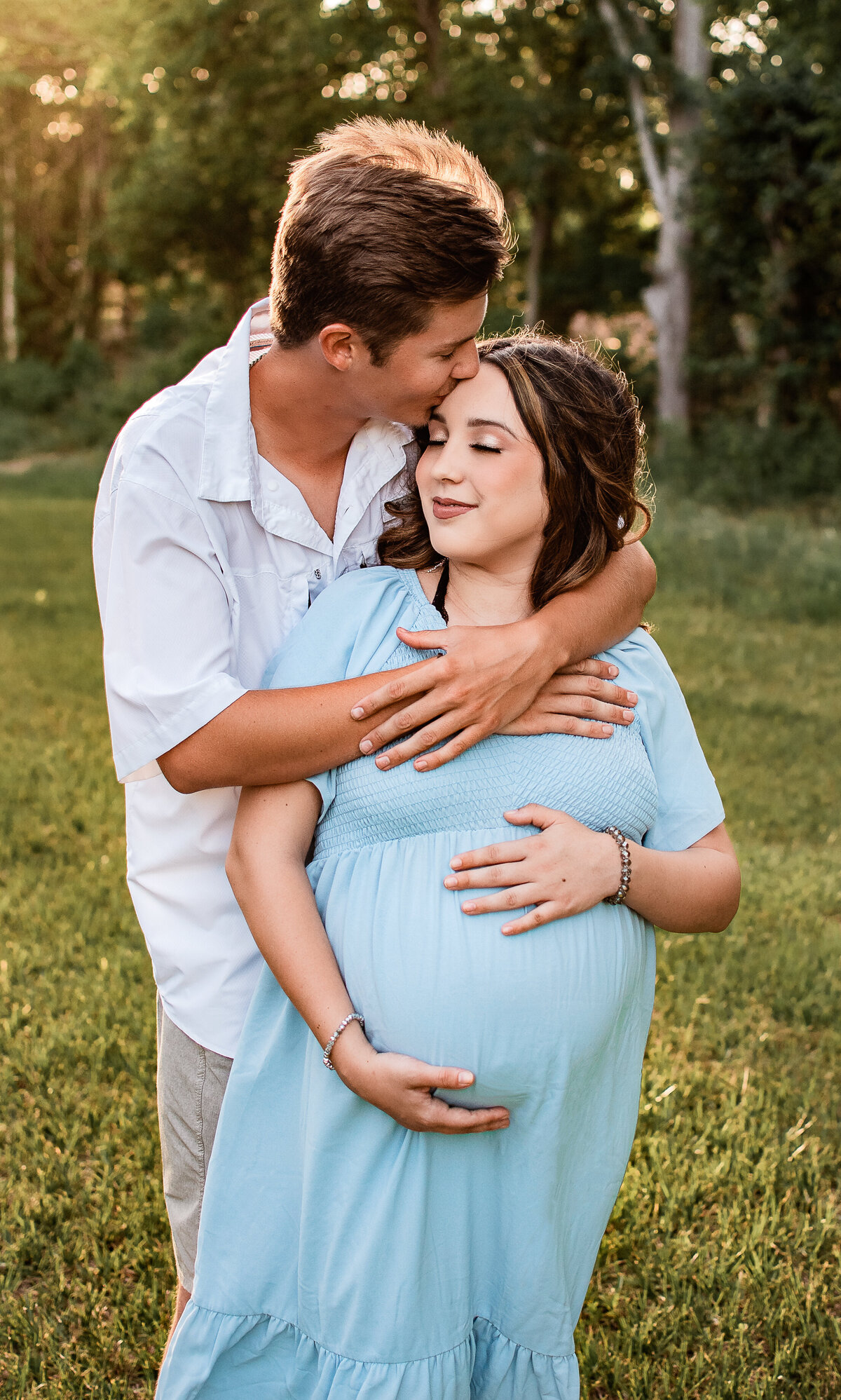 An expectant dad hugs his pregnant girlfriend around the neck while she touches her belly.