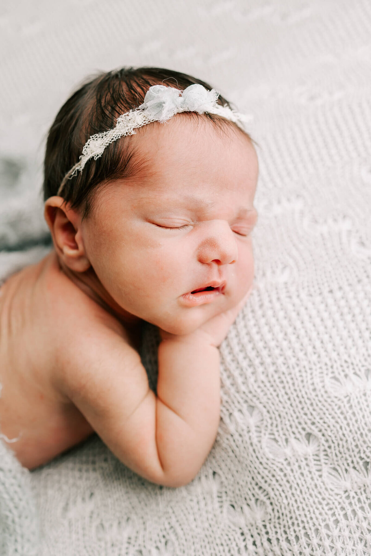 infant girl sleeping on a grey blanket with her hands under her chin. she is wearing a blue bow headband
