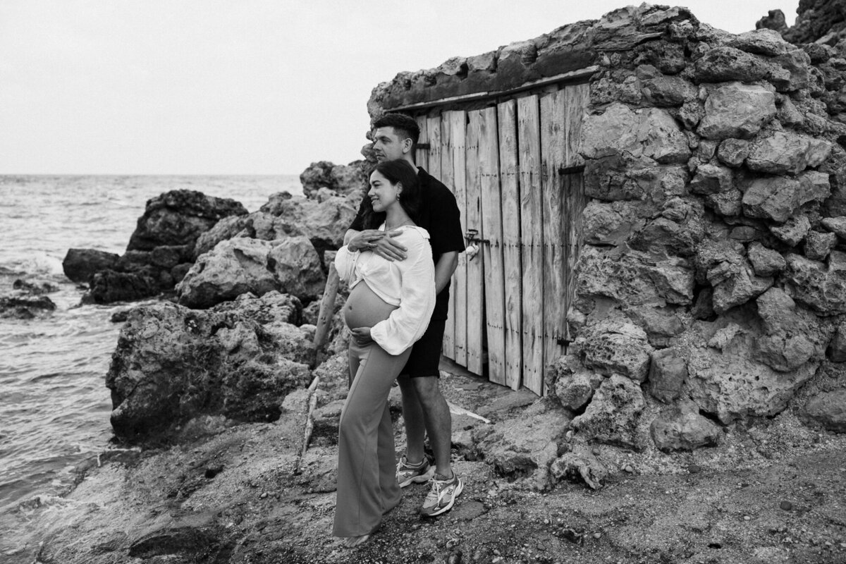 Man standing behind a woman in front of a fisherman's cabin on Ibiza for a pregnancy photography