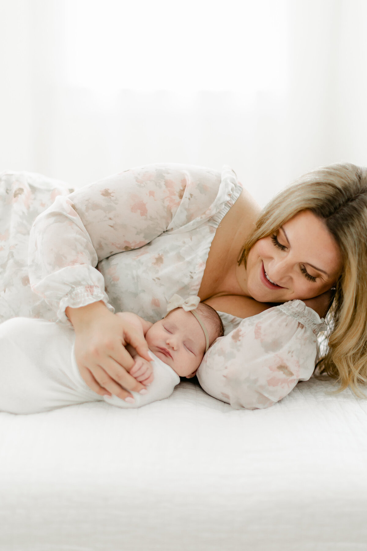 new mom wearing a beautiful floral dress laying on a white bed with her newborn baby girl photographed by Philadelphia Newborn Photographer Tara Federico