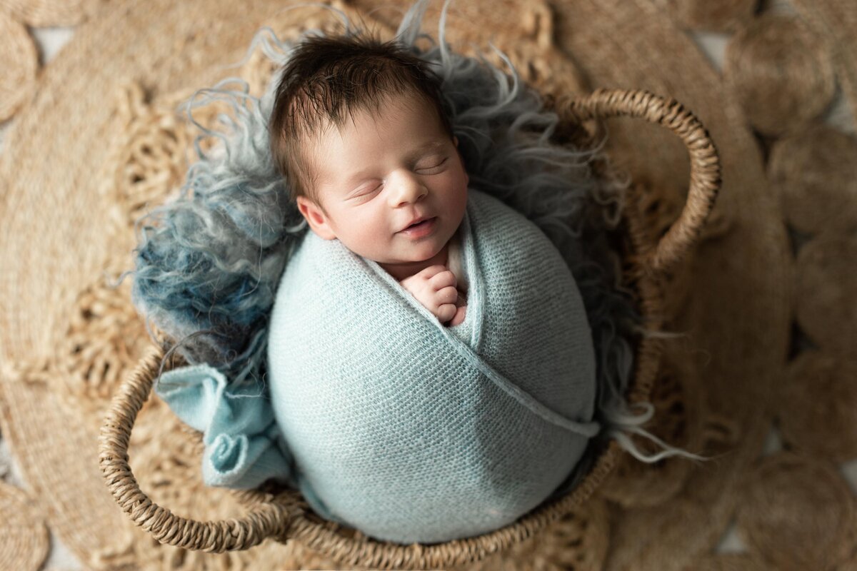 Studio newborn photography - baby sleeping in a woven basket on top of a large, boho inspired woven mat. Baby is wrapped in a seafoam knit swaddle with their hands peeking out of the wrap and slightly smiling.