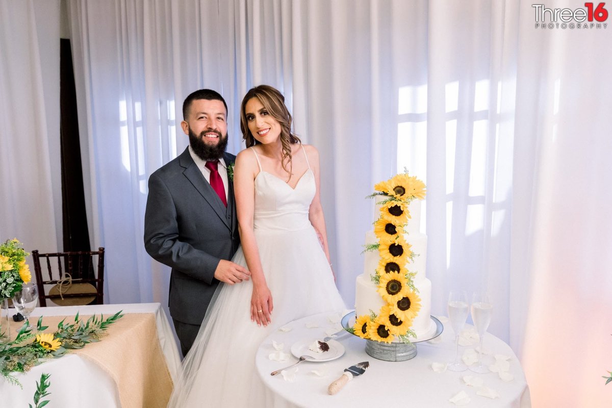 Bride and Groom pose next to their three-tiered wedding cake draped by sunflowers