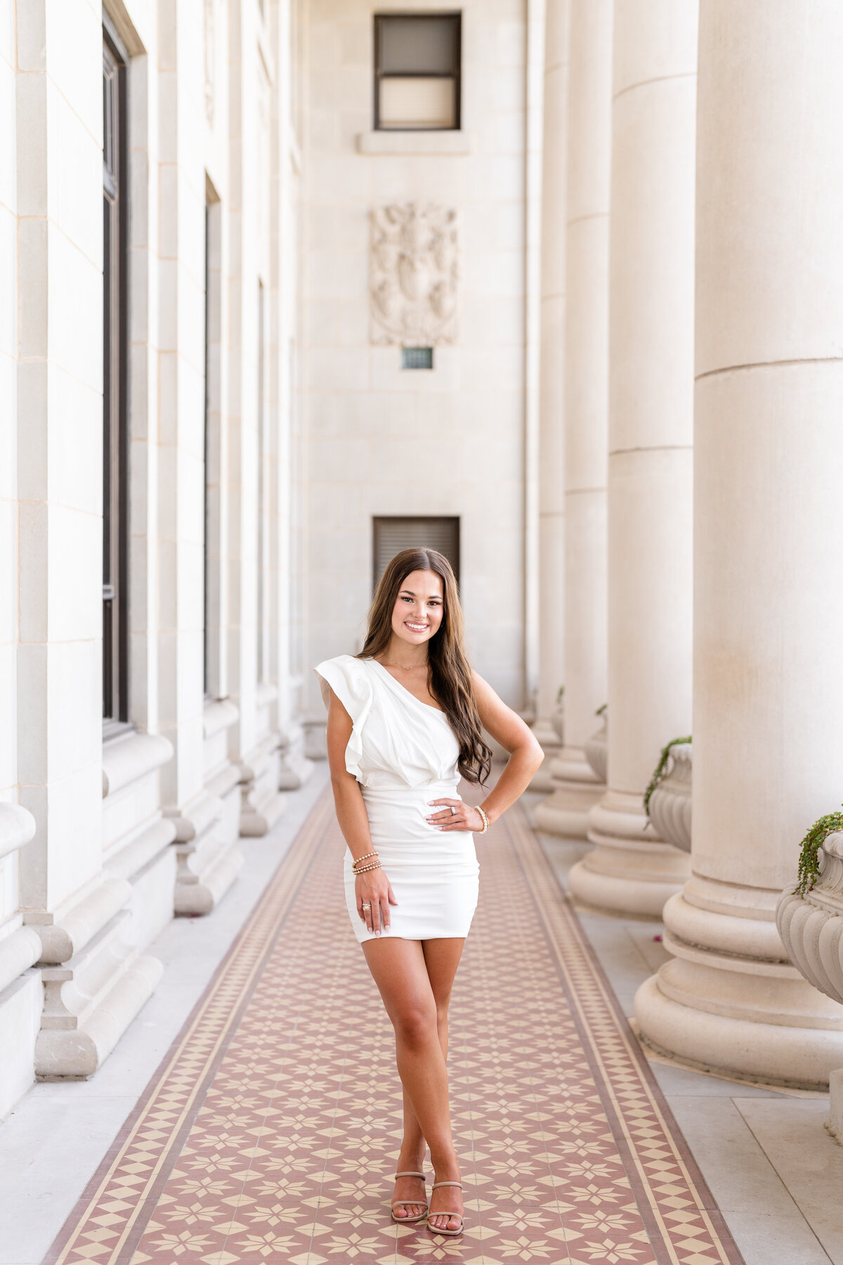 Texas A&M senior girl smiling with hand on hip and wearing white one shoulder dress in the columns of the Administration Building