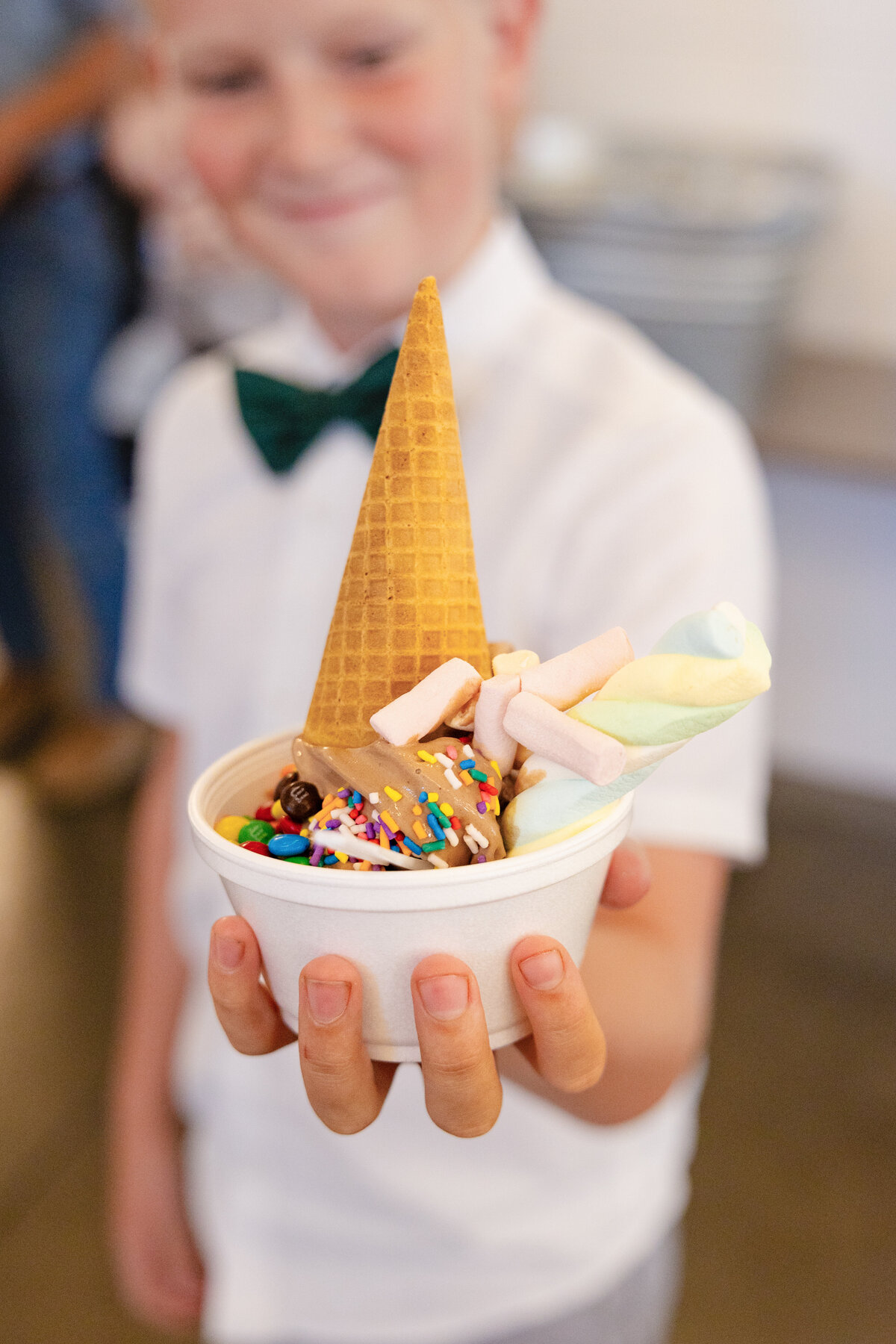 A young boy in a bowtie holds a cup of ice cream topped with sprinkles, marshmallows, and an upside-down cone.