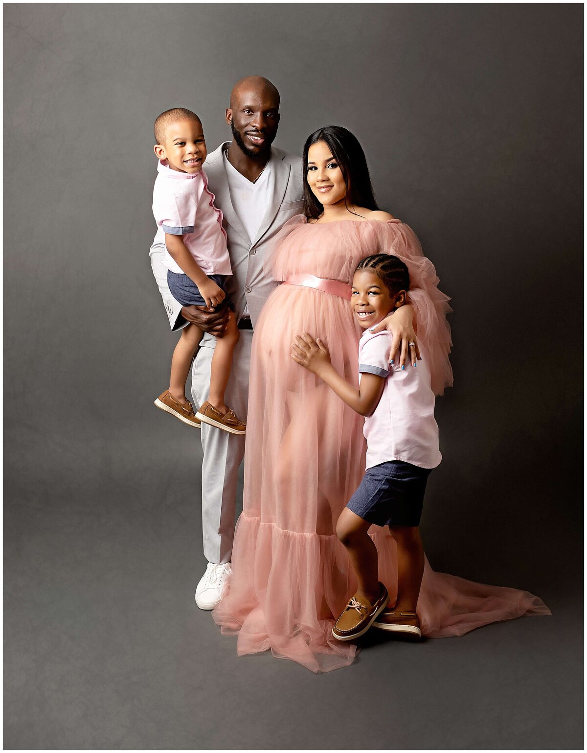A family of four poses in studio for a pregnancy photoshoot, with the father holding up the younger child and the older child standing and hugging moms belly. The photo captures the anticipation and happiness of the growing family as they prepare to welcome a new addition.