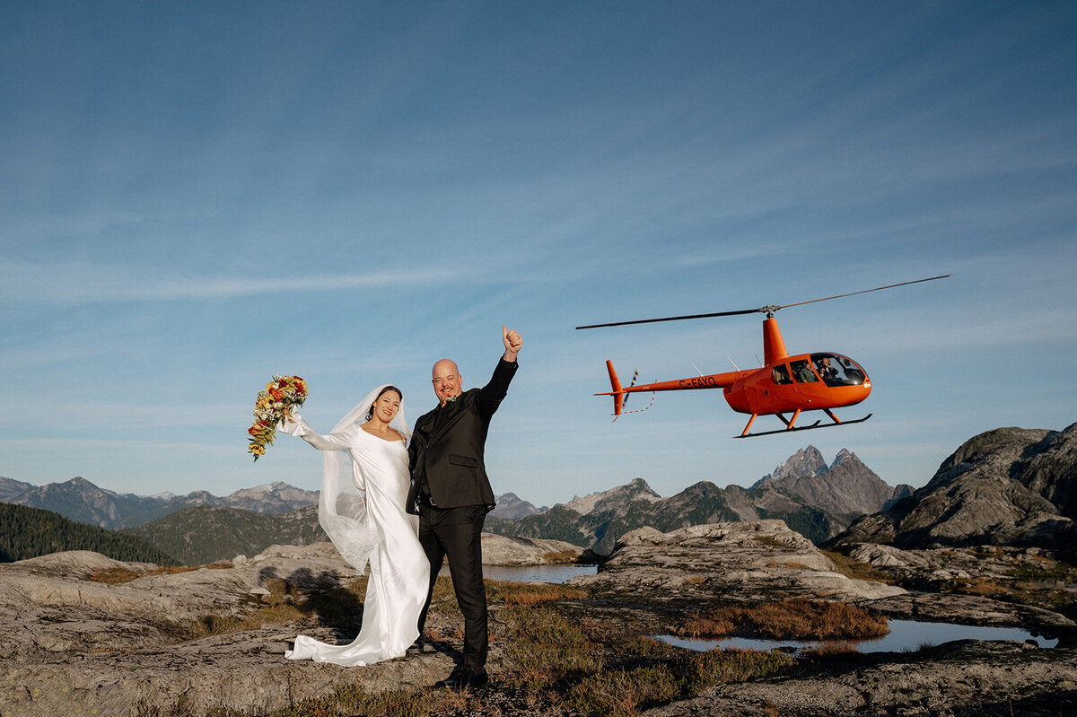 Bride and groom in front of an airborne orange helicopter after being dropped off to take a photo