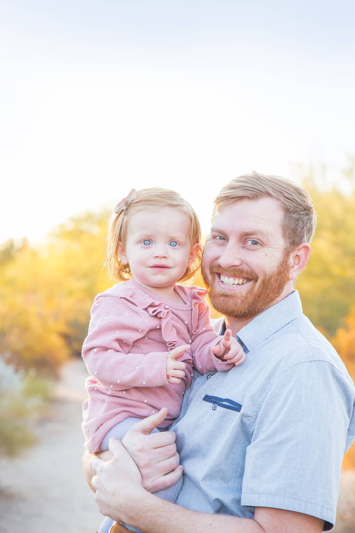 Toddler girl in pink dress with red strawberry blonde hair held by smiling red haired dad in desert park