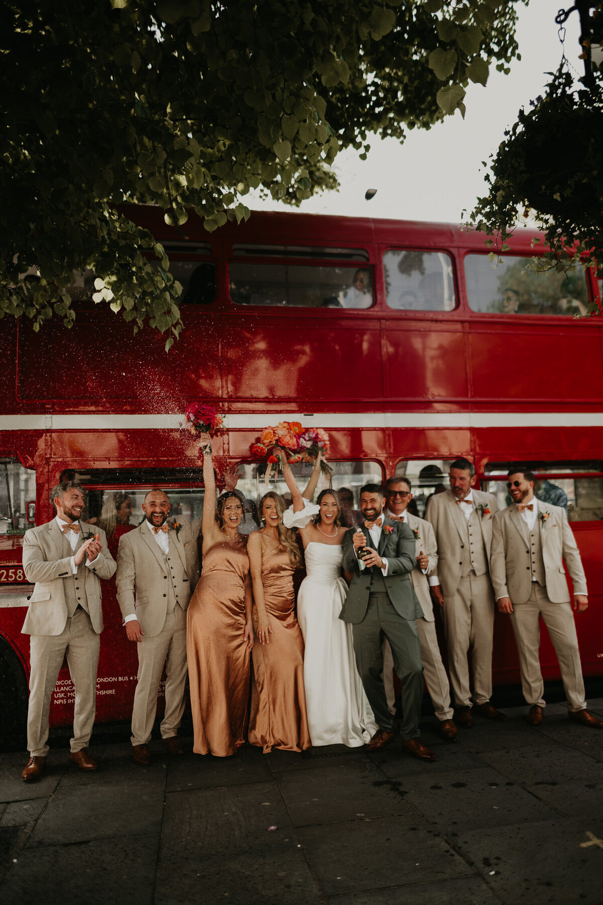A bride and groom along with their wedding party spray champagne in front of a red London bus. 