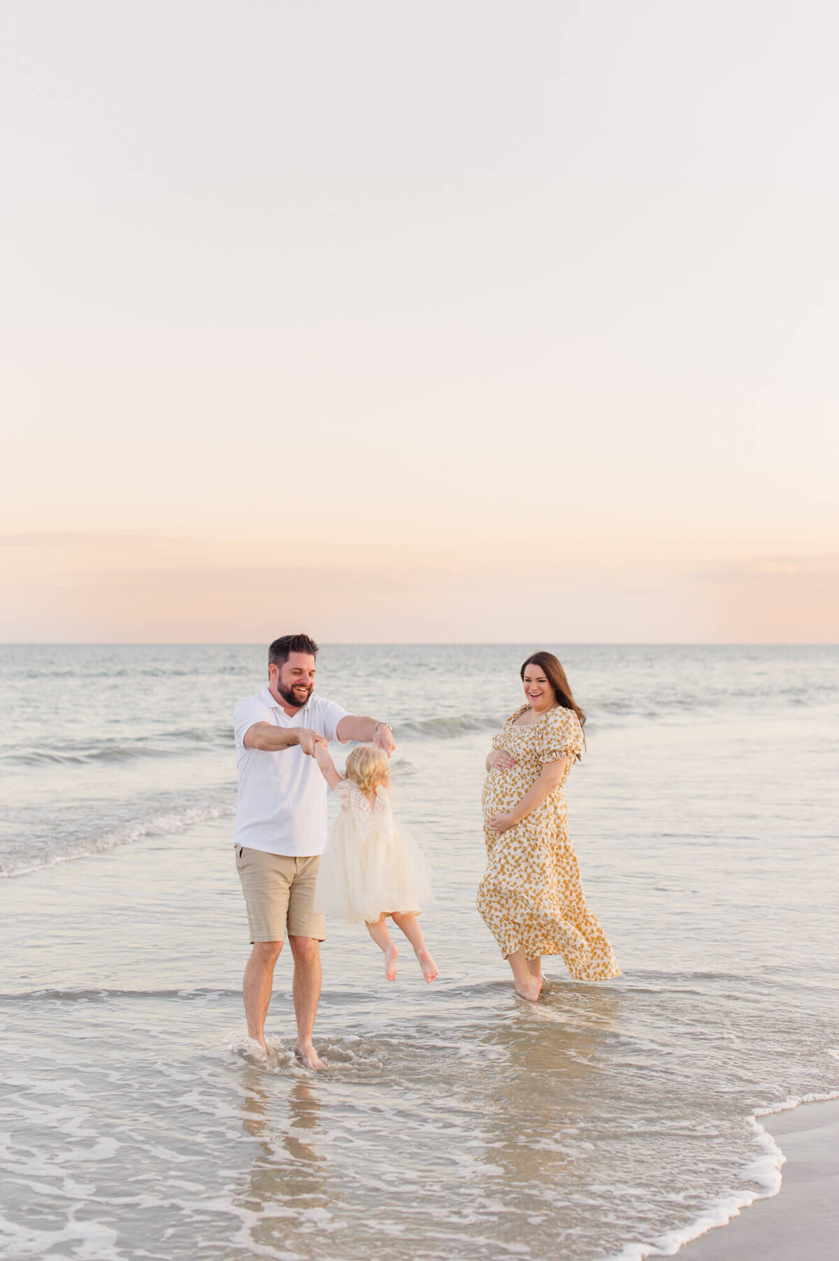 Orlando maternity photographer captures expectant parents playing in the ocean waves with their young toddler girl at sunset