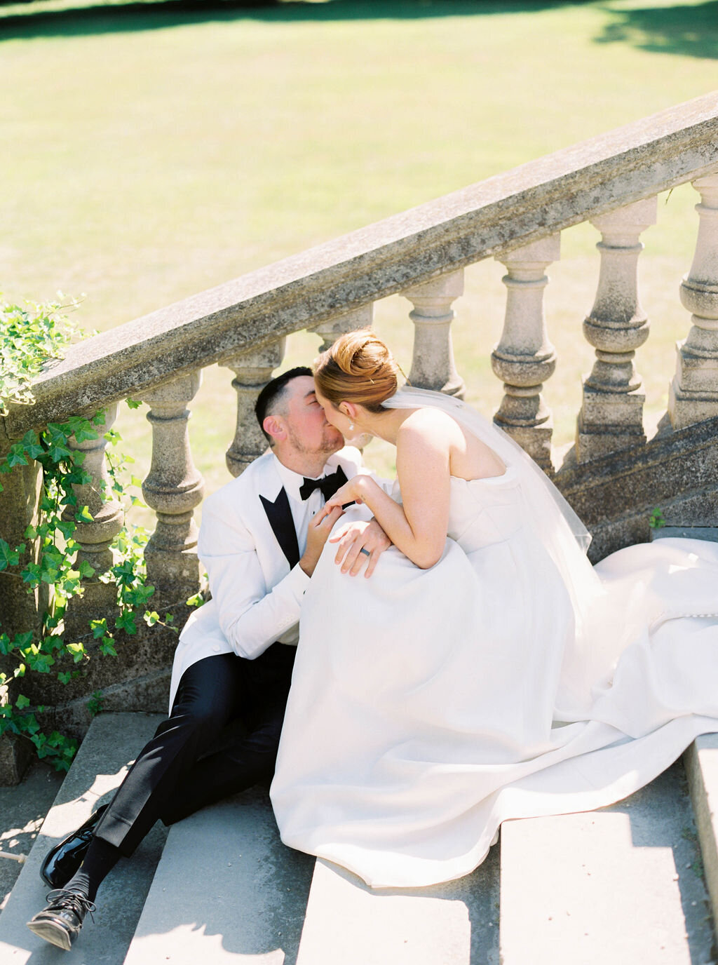 Rhode Island Elopement | A couple sits on stone steps, sharing a tender moment. The bride in a flowing white dress and groom in a white tuxedo kiss beside an ivy-clad balustrade.