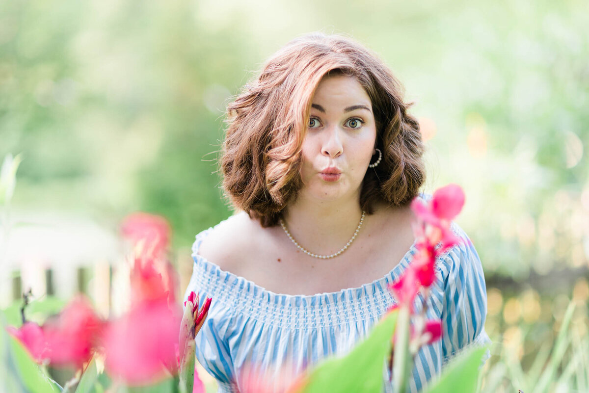 Fun senior portrait of a girl in a blue and white shirt blowing a kiss at the camera behind pink flowers.