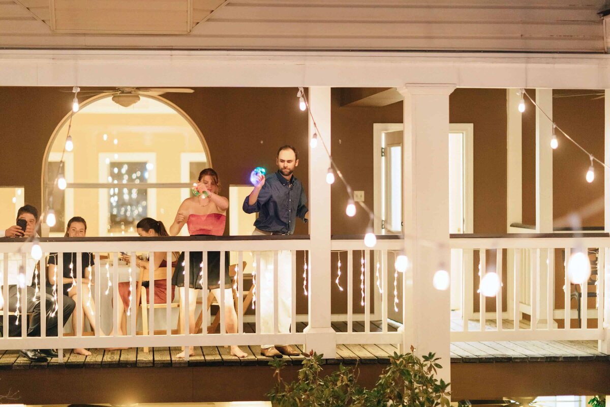A group of people stand on a balcony with white railings decorated with string lights at night; a man holds a lit-up ball while a woman beside him blows bubbles, and others sit or take photos in a relaxed, festive atmosphere.